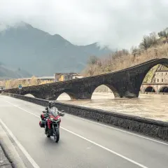 Motociclista attraversa il Ponte del Diavolo, un viaggio tra Lucca e Siena, con il fiume in piena sotto le arcate.