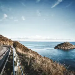 Un motociclista percorre una strada panoramica sull'Argentario, con il mare blu e un'isola rocciosa sullo sfondo.