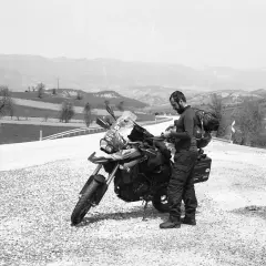 Motociclista con zaino e attrezzatura da viaggio si ferma accanto alla sua moto, la sella ciampigotto pronta per la prossima avventura. Paesaggio collinare sullo sfondo.