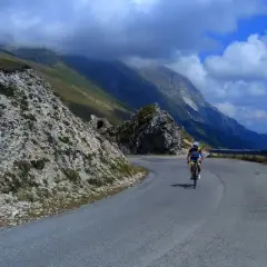 Ciclista in salita sulla strada panoramica della Forca di Presta, con montagne sullo sfondo e una moto parcheggiata.