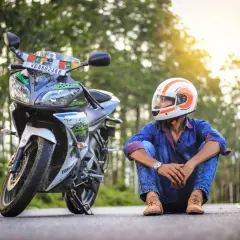 Giovane con casco arancione e bianco, seduto accanto alla sua moto. Spera di allargare il casco stretto per un viaggio più comodo.