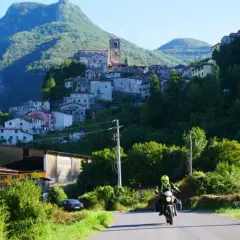 Un motociclista esplora la Lunigiana, con un borgo arroccato su una collina verdeggiante sullo sfondo.