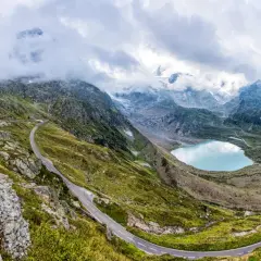 Strada tortuosa sul passo della Sambuca, con lago alpino azzurro e montagne innevate sullo sfondo.