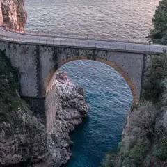 Ponte ad arco in pietra che attraversa un fiordo, con il mare blu sottostante. Un'icona del furore costiera amalfitana, incastonata tra rocce e vegetazione.