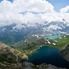 Strada tortuosa sul passo Nivolet, con laghi alpini e montagne innevate sotto un cielo nuvoloso.