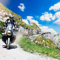 Motociclista affronta un ripido tornante della boaria su una strada sterrata in montagna, con un cielo azzurro e nuvole bianche.