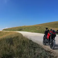Ducati rossa ferma su strada sterrata, con un passo branchetto sullo sfondo e cielo azzurro.