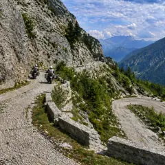 Due motociclisti affrontano tornanti mozzafiato sul passo del Tarvisio, tra rocce imponenti e valli verdi.