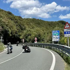 Motociclisti in curva sul passo del Bracco, tra il verde della Liguria e un cielo azzurro con nuvole bianche.