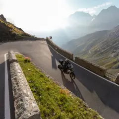 Motociclista affronta tornanti mozzafiato sul Passo Giovo, con montagne maestose e raggi di sole che illuminano il paesaggio alpino.