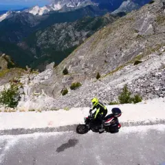 Un motociclista con casco giallo affronta il passo del vestito in moto, tra montagne rocciose e cave di marmo.