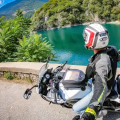Weekend in moto nel Nord Italia: un motociclista ammira un lago alpino turchese, con montagne verdi sullo sfondo.