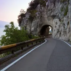 Strada della forra in moto, con un tunnel scavato nella roccia e il lago sullo sfondo al tramonto.