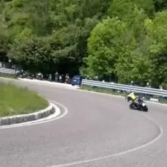 Un motociclista in giallo affronta una curva su una **strada del costo** panoramica, con spettatori e moto parcheggiate a lato.