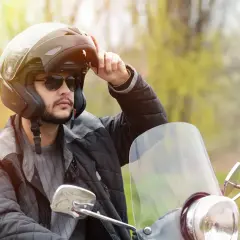 Uomo con casco da moto, occhiali da sole e giacca imbottita, che regola le parti del casco.