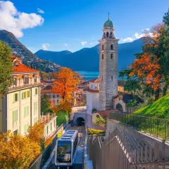 Un trenino scende tra case colorate e un campanile, con il Lago Maggiore sullo sfondo. Un assaggio delle **strade panoramiche Lago Maggiore** in autunno.