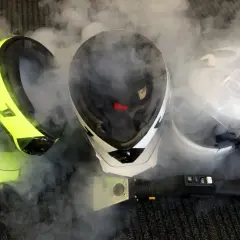 Tre caschi, uno giallo fluo, uno bianco e nero e uno bianco, circondati da vapore. Un pulitore a vapore è posizionato davanti, suggerendo che il casco non deve essere pulito con solventi.