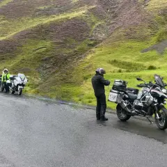 Motociclisti in sosta sul passo della Contessa, con moto cariche e un paesaggio collinare verde e viola sullo sfondo.