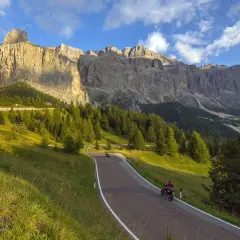 Strada tortuosa tra prati verdi e boschi di conifere, con imponenti vette dolomitiche sullo sfondo. Due motociclisti godono del panorama mozzafiato, un'avventura indimenticabile tra le Dolomiti in moto.