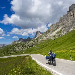 Motociclista percorre la strada cimina tra prati verdi e montagne imponenti sotto un cielo azzurro con nuvole bianche.