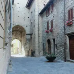 Un vicolo suggestivo a Varzi, con un arco in pietra che conduce a una strada illuminata. Case antiche e fiori colorati invitano a scoprire cosa vedere.