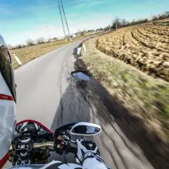 In sella a una moto, con il casco bianco e rosso, si percorre una strada di campagna. Il campo arato a lato e il cielo azzurro evocano il linguaggio motociclisti italiano.