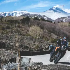 Motociclista su strada mareneve, con vulcano innevato sullo sfondo.