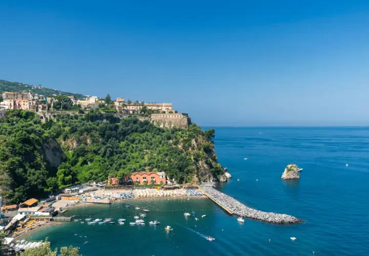 Vico Equense cosa vedere: una baia con spiaggia attrezzata, un molo di rocce e un borgo arroccato sulla scogliera.