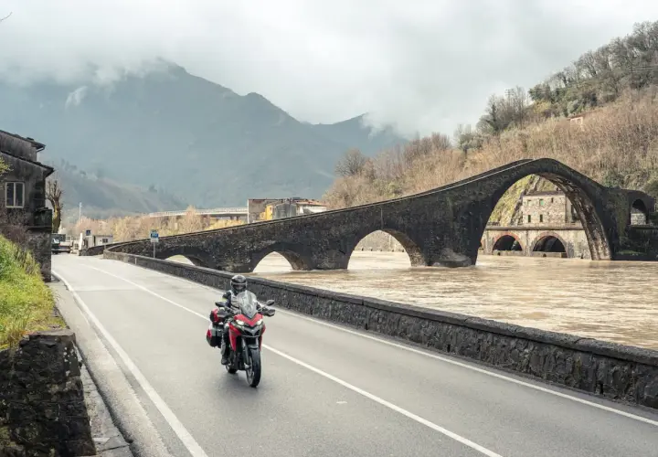 Motociclista attraversa il Ponte del Diavolo, un viaggio tra Lucca e Siena, con il fiume in piena sotto le arcate.