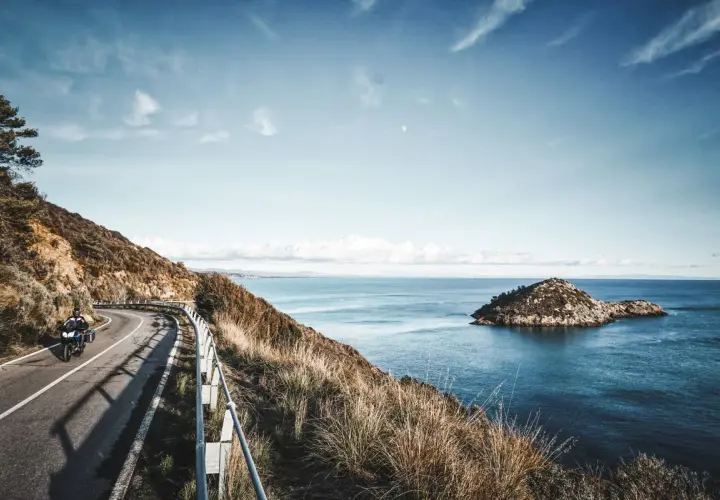 Un motociclista percorre una strada panoramica sull'Argentario, con il mare blu e un'isola rocciosa sullo sfondo.