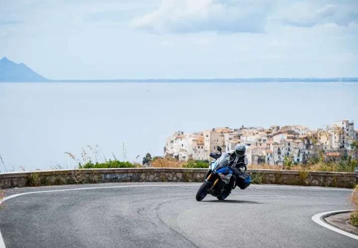 Motociclista in curva su strada panoramica, con il mare e il borgo di Sperlonga sullo sfondo. Un viaggio da Terracina a Sperlonga.
