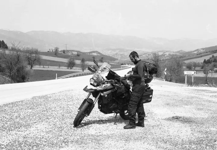 Motociclista con zaino e attrezzatura da viaggio si ferma accanto alla sua moto, la sella ciampigotto pronta per la prossima avventura. Paesaggio collinare sullo sfondo.