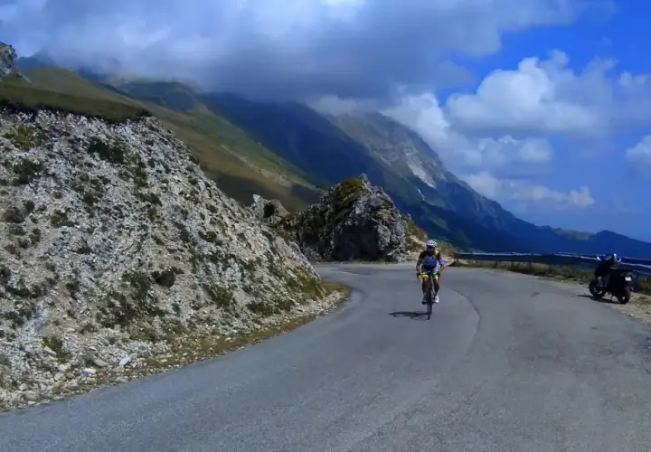 Ciclista in salita sulla strada panoramica della Forca di Presta, con montagne sullo sfondo e una moto parcheggiata.