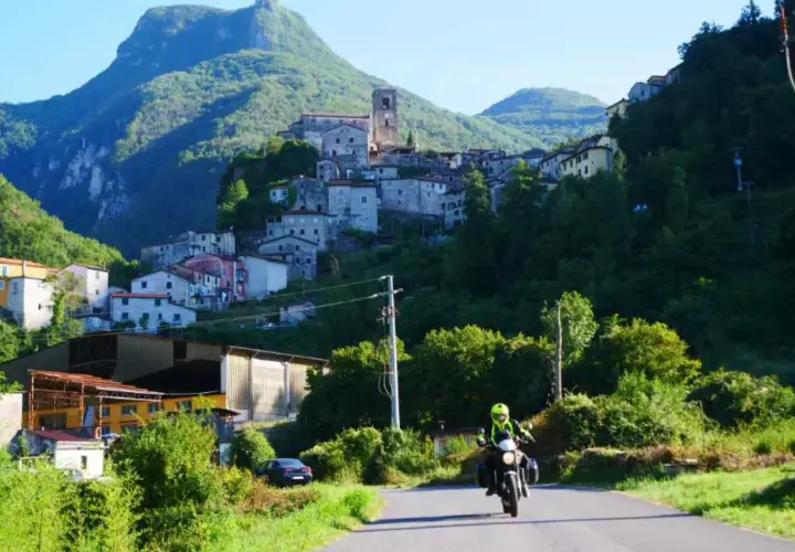 Un motociclista esplora la Lunigiana, con un borgo arroccato su una collina verdeggiante sullo sfondo.