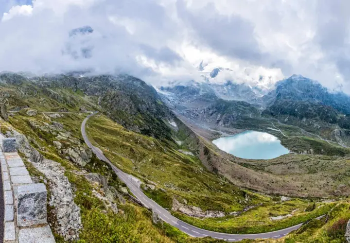 Strada tortuosa sul passo della Sambuca, con lago alpino azzurro e montagne innevate sullo sfondo.