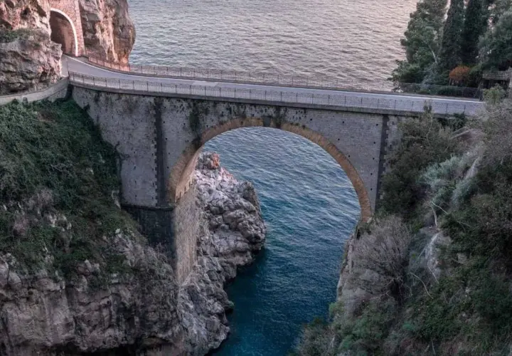 Ponte ad arco in pietra che attraversa un fiordo, con il mare blu sottostante. Un'icona del furore costiera amalfitana, incastonata tra rocce e vegetazione.