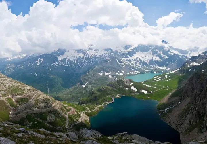 Strada tortuosa sul passo Nivolet, con laghi alpini e montagne innevate sotto un cielo nuvoloso.