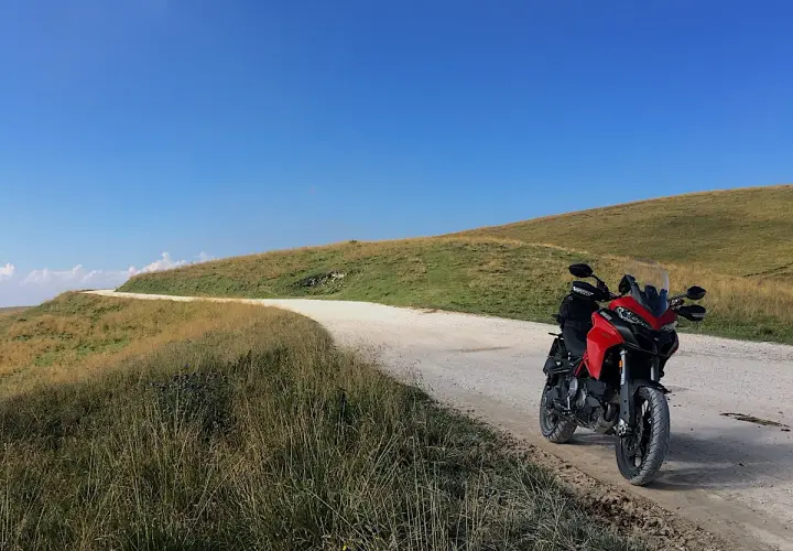 Ducati rossa ferma su strada sterrata, con un passo branchetto sullo sfondo e cielo azzurro.