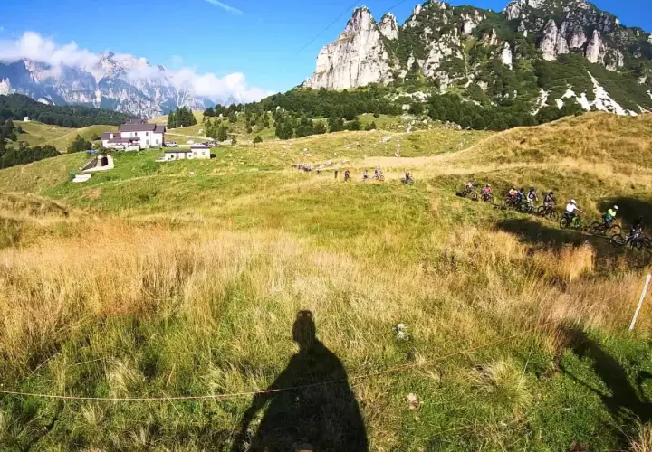 Gruppo di ciclisti in discesa su un sentiero erboso, con montagne imponenti sullo sfondo. Un'avventura a Recoaro 1000.