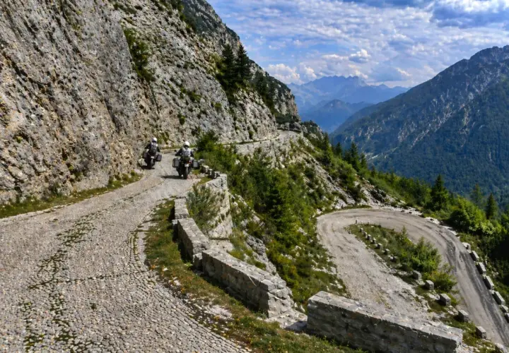 Due motociclisti affrontano tornanti mozzafiato sul passo del Tarvisio, tra rocce imponenti e valli verdi.