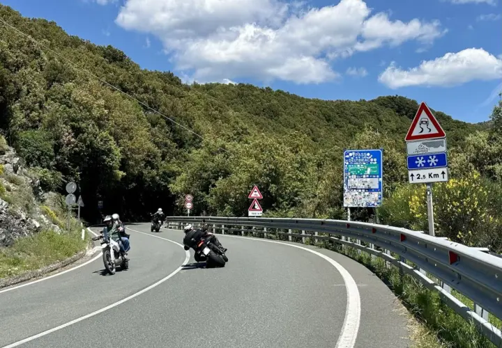 Motociclisti in curva sul passo del Bracco, tra il verde della Liguria e un cielo azzurro con nuvole bianche.