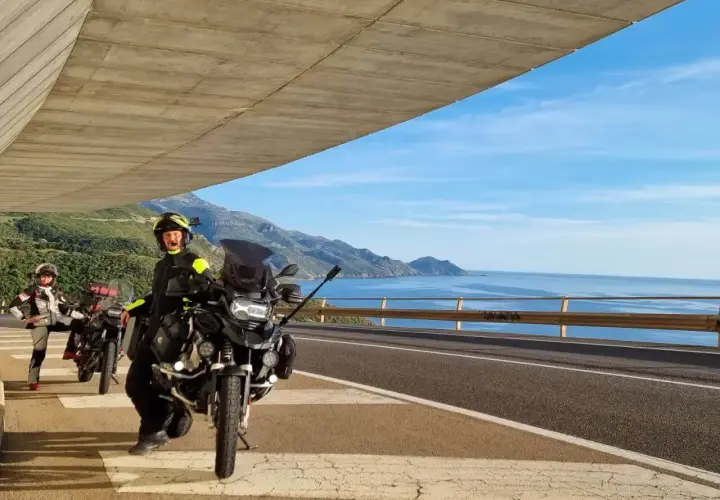 Motociclisti in sosta panoramica, con moto pronte per esplorare la costa tra Alghero e Oristano. Mare azzurro e montagne sullo sfondo.