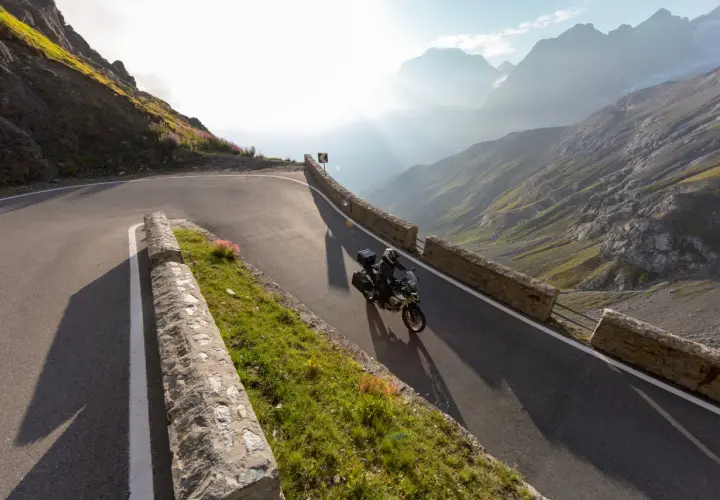 Motociclista affronta tornanti mozzafiato sul Passo Giovo, con montagne maestose e raggi di sole che illuminano il paesaggio alpino.