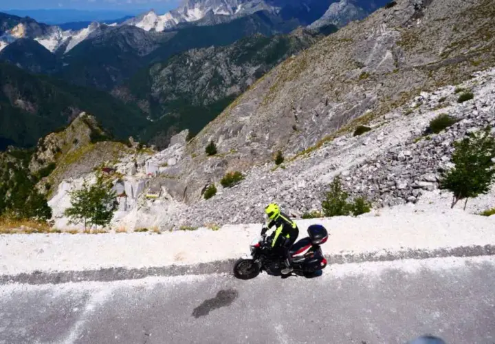 Un motociclista con casco giallo affronta il passo del vestito in moto, tra montagne rocciose e cave di marmo.