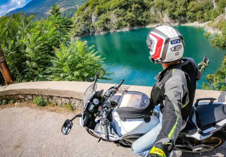 Weekend in moto nel Nord Italia: un motociclista ammira un lago alpino turchese, con montagne verdi sullo sfondo.