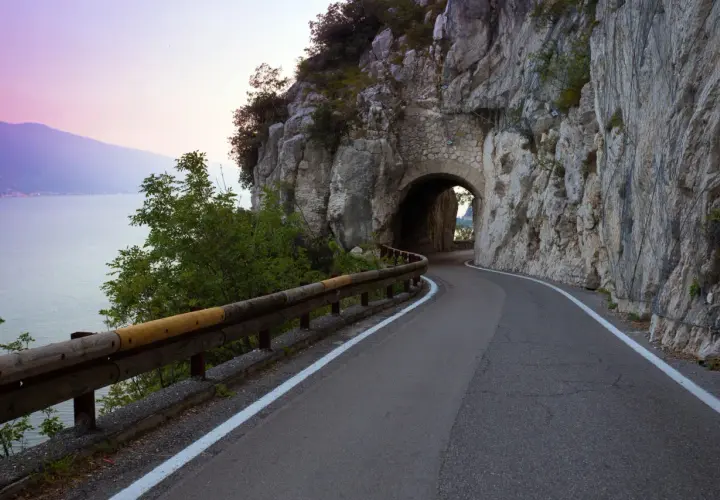 Strada della forra in moto, con un tunnel scavato nella roccia e il lago sullo sfondo al tramonto.
