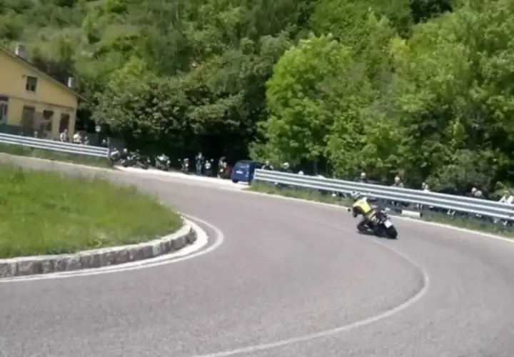 Un motociclista in giallo affronta una curva su una **strada del costo** panoramica, con spettatori e moto parcheggiate a lato.
