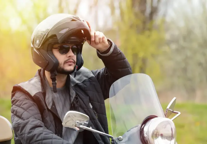 Uomo con casco da moto, occhiali da sole e giacca imbottita, che regola le parti del casco.