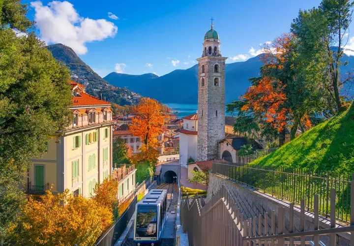 Un trenino scende tra case colorate e un campanile, con il Lago Maggiore sullo sfondo. Un assaggio delle **strade panoramiche Lago Maggiore** in autunno.