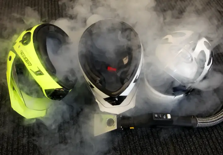 Tre caschi, uno giallo fluo, uno bianco e nero e uno bianco, circondati da vapore. Un pulitore a vapore è posizionato davanti, suggerendo che il casco non deve essere pulito con solventi.